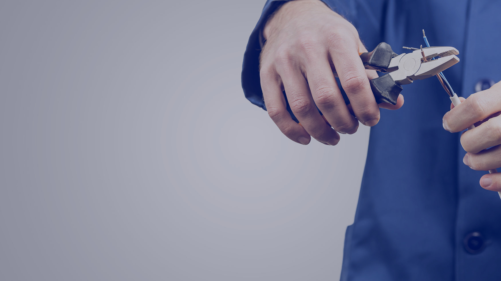 Workman or electrician repairing an electrical cable with a pair of pliers to restore supply to the house, close up view of his hands in blue overalls on grey with copyspace